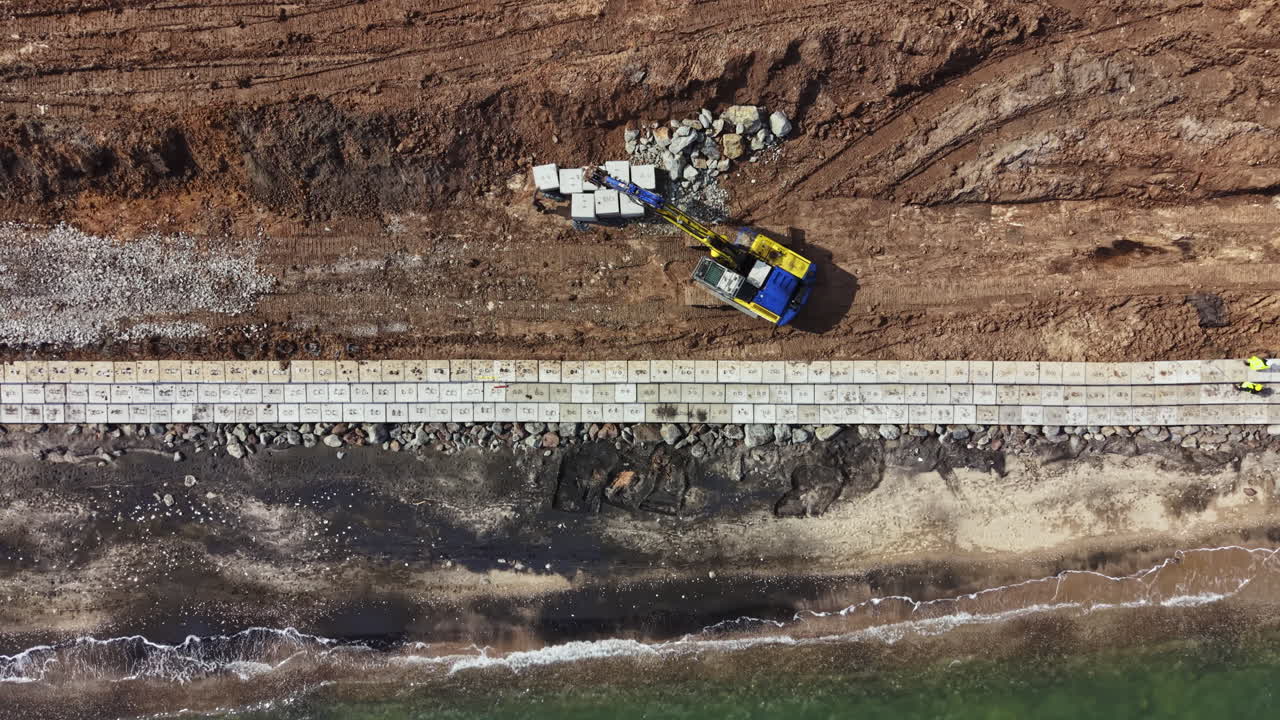Lavori di costruzione in un'area costiera con macchinari che spostano materiali vicino al litorale durante le ore diurne.