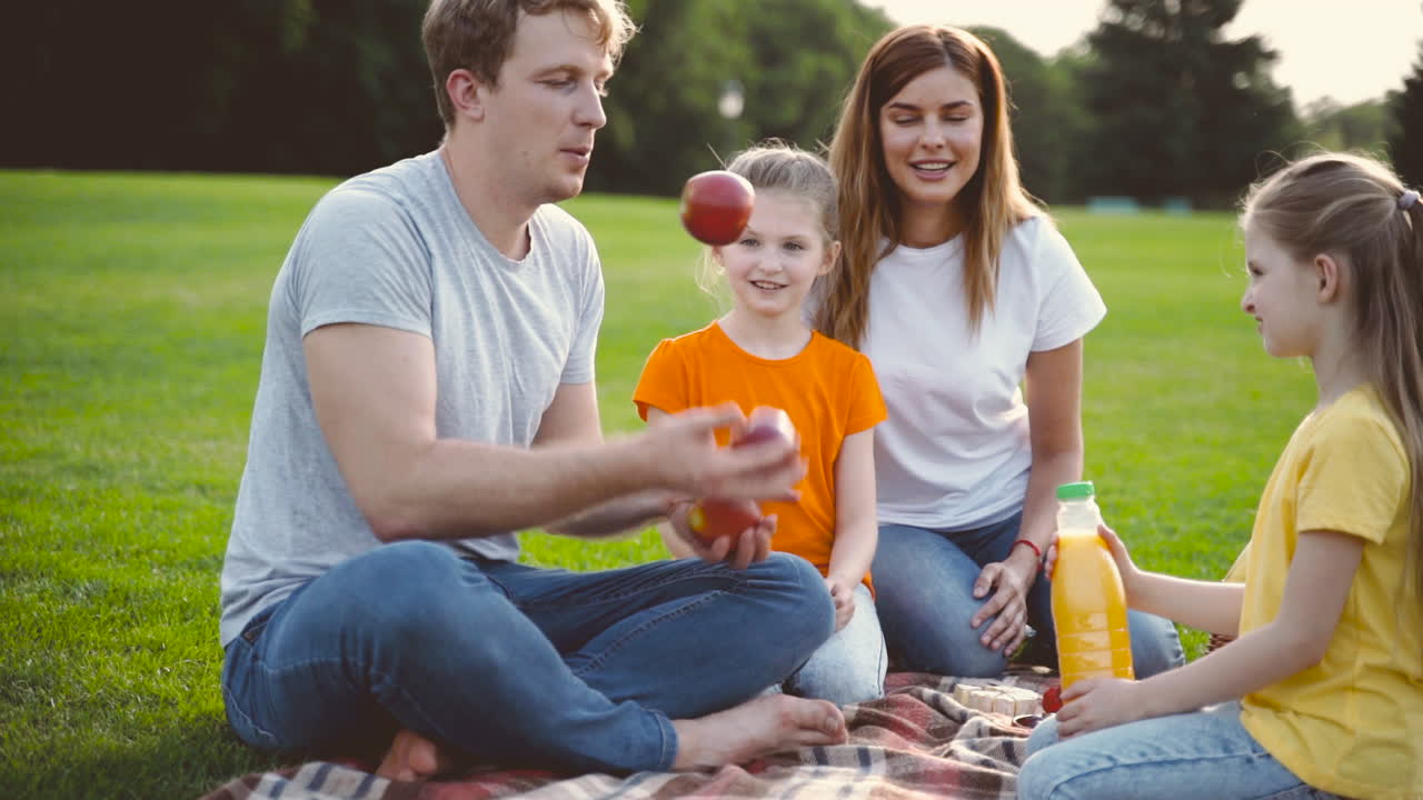 padre haciendo malabarismos con manzanas y divirtiéndose con su esposa y sus dos hijas pequeñas durante un picnic en un prado verde en el parque