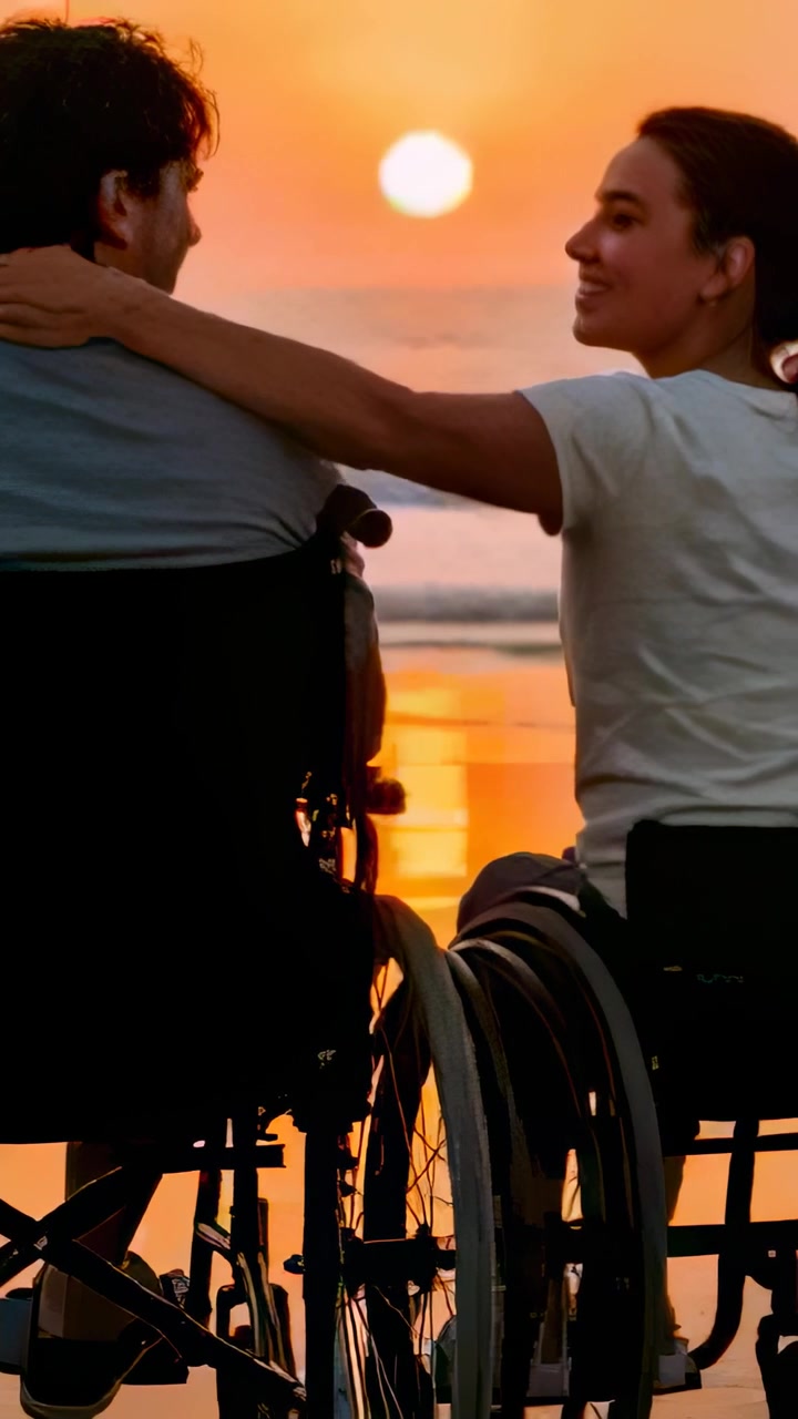 Vertical video: Sun sinking, right partner reaching, holding hands at beach in manual wheelchairs