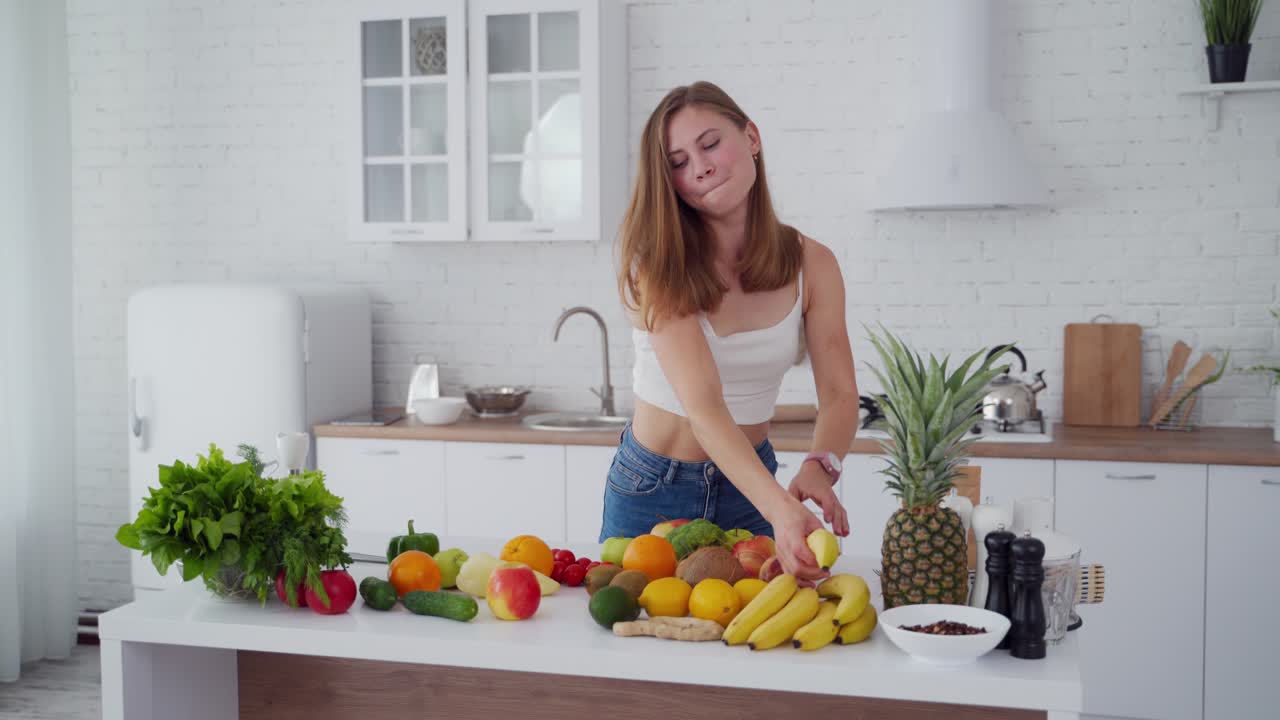 Happy young female in the kitchen. Beautiful lady throws away centimeter and takes banana. Fresh fruit and vegetables on the table. Healthy diet.