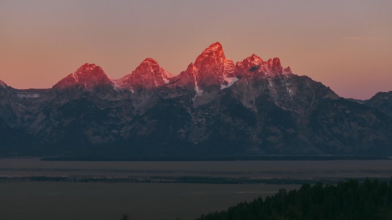 Sunrise light illuminating snow capped Grand Teton mountain range above valley, aerial backdrop alpenglow at sunset golden hour