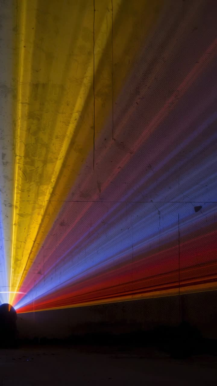 Vídeo vertical: projeção de uma pequena luminária que emite feixes amarelos, azuis e vermelhos no teto de um túnel com névoa