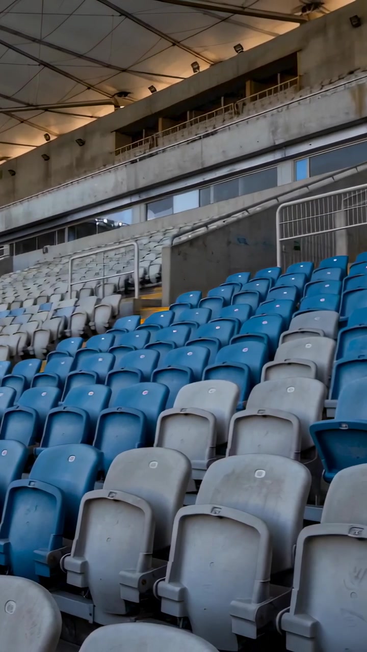 Video vertical: Cámara panorámica revelando asientos vacíos de color azul grisáceo en un estadio para mostrar el palco de prensa y la barandilla.