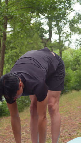video vertical de un hombre con ropa deportiva haciendo yoga se estira en una alfombra en el bosque junto a un lago o río disfrutando de la paz y la belleza de la naturaleza filmado en tiempo real 1