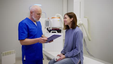 Confident-male-doctor-with-a-gray-beard-in-glasses-and-in-a-blue-uniform-interviews-and-listens-to-the-complaints-of-a-brunette-girl-during-an-appointment-with-a-doctor-in-a-modern-clinic