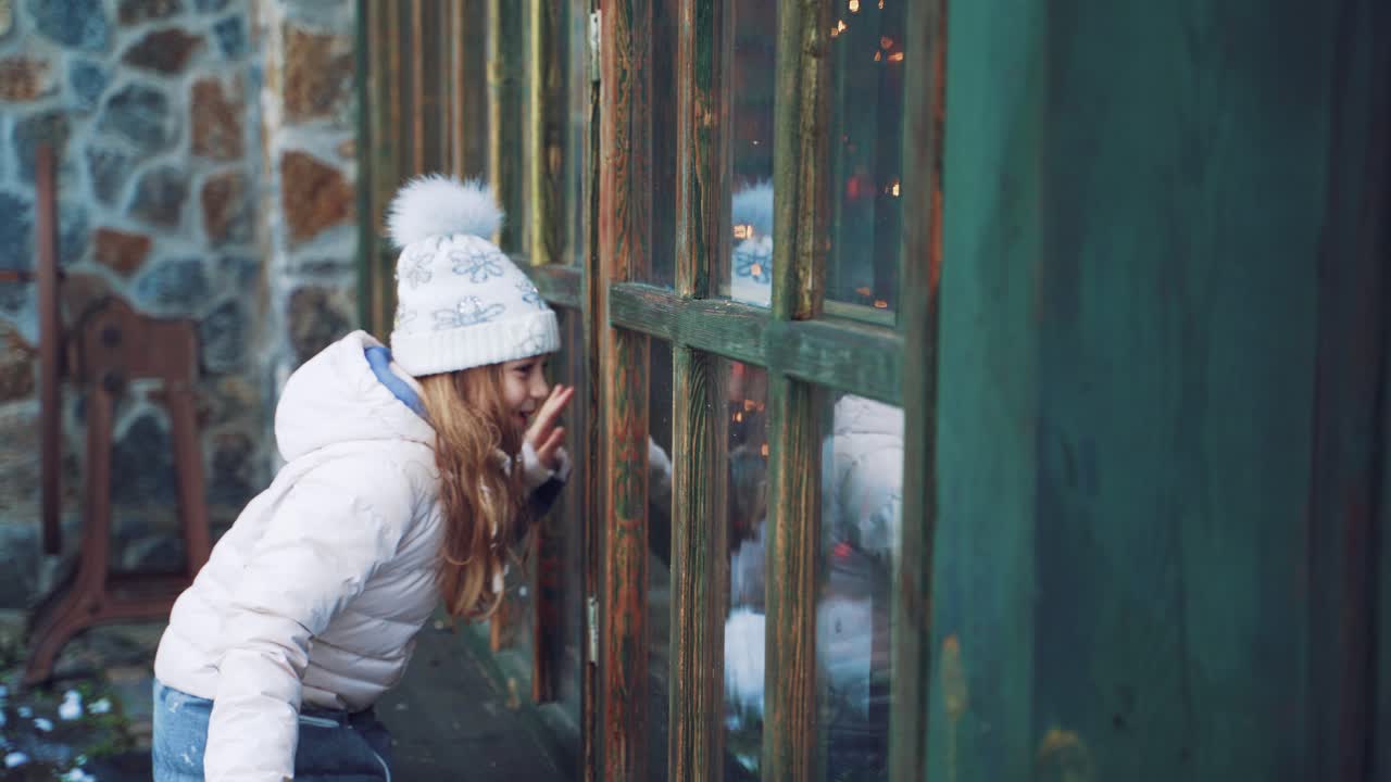 Cheerful little girl with long hair and in warm clothes is peeping through the window from the street behind the elves in the winter against the background of the building. Good New Year spirit