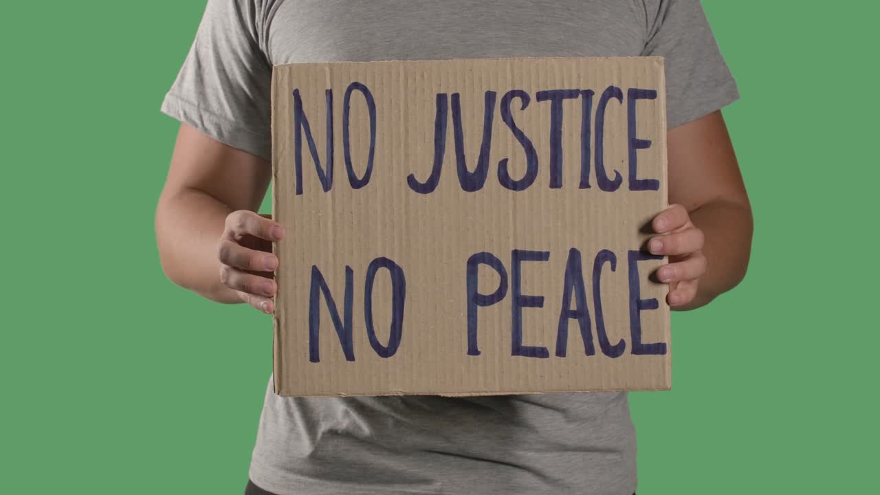"그의 앞에는 ""no justice, no peace""라는 문구가 새겨진 카드보드 상자에 붙어 있는 포스터가 붙어 있다.그는 파업과 반 인종차별의 개념, 녹색 스크린, 크로마 키, 클로즈업, 느린 동작을 고립시킨다."