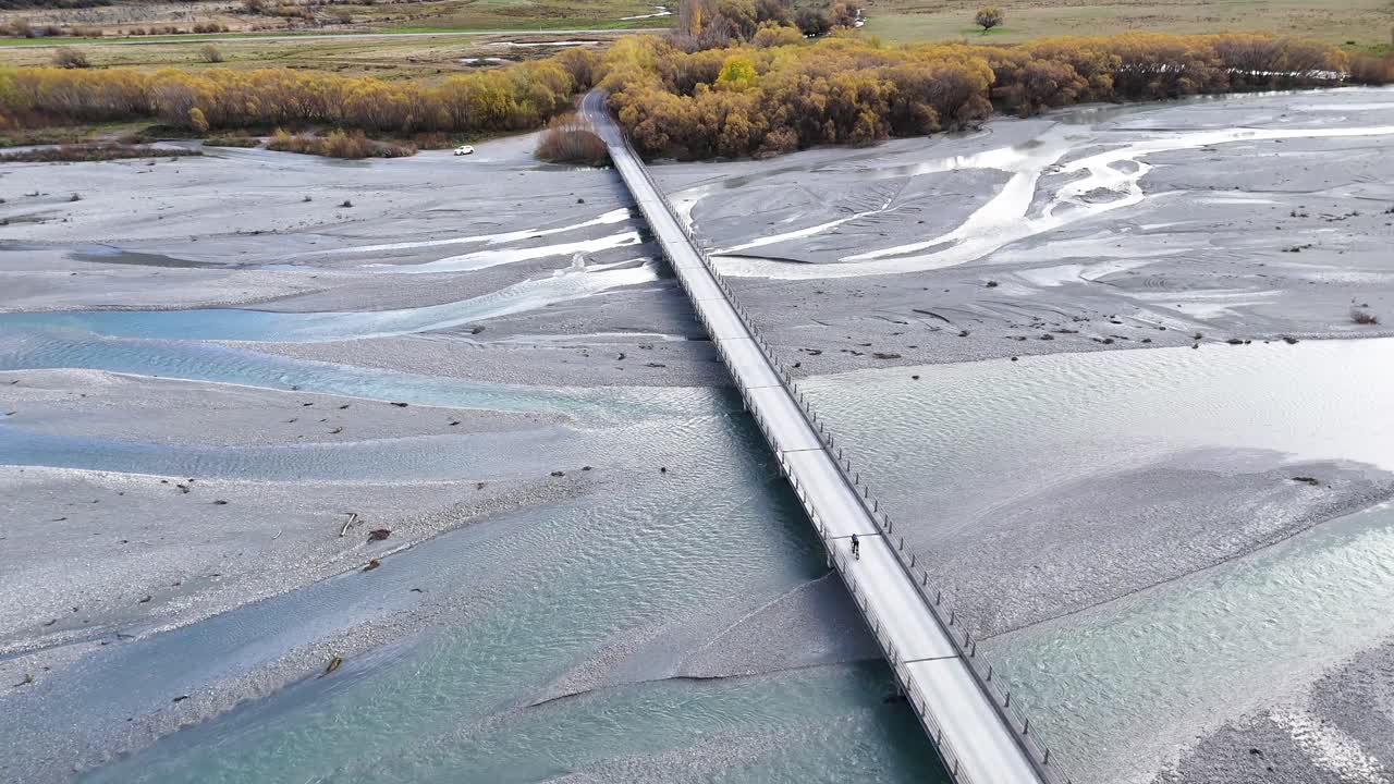 A cyclist rides across a long bridge over a braided river in Glenorchy, New Zealand. Bright daylight enhances the vibrant scenery