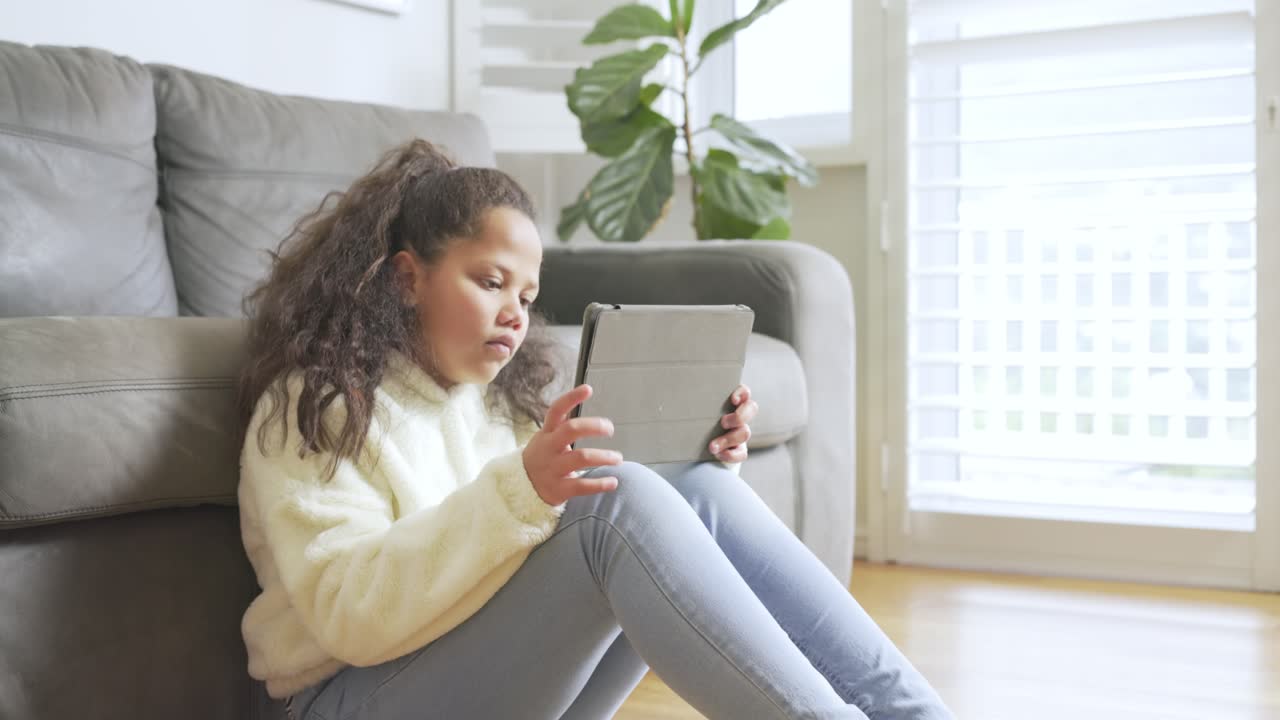 School-age girl sitting on floor by sofa tapping tablet screen, swiping to solve puzzle, copy space