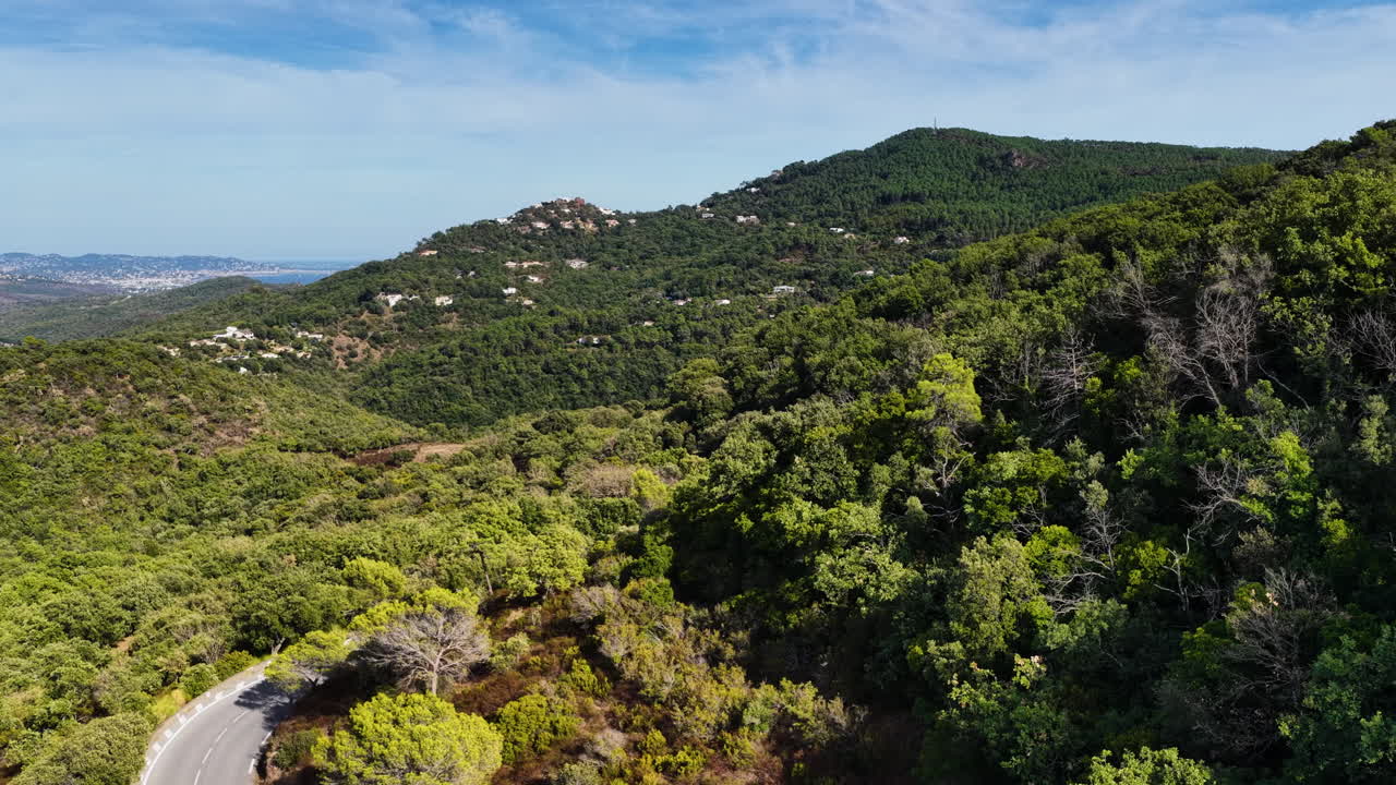 Veduta aerea con drone di una strada tortuosa attraverso verdi colline mediterranee, con lo skyline di Monaco visibile in lontananza lungo la Costa Azzurra.