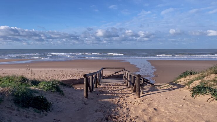 Ein hölzerner Bohlenweg führt durch die Küstenvegetation zum Atlantikstrand unter einem klaren, blauen Himmel