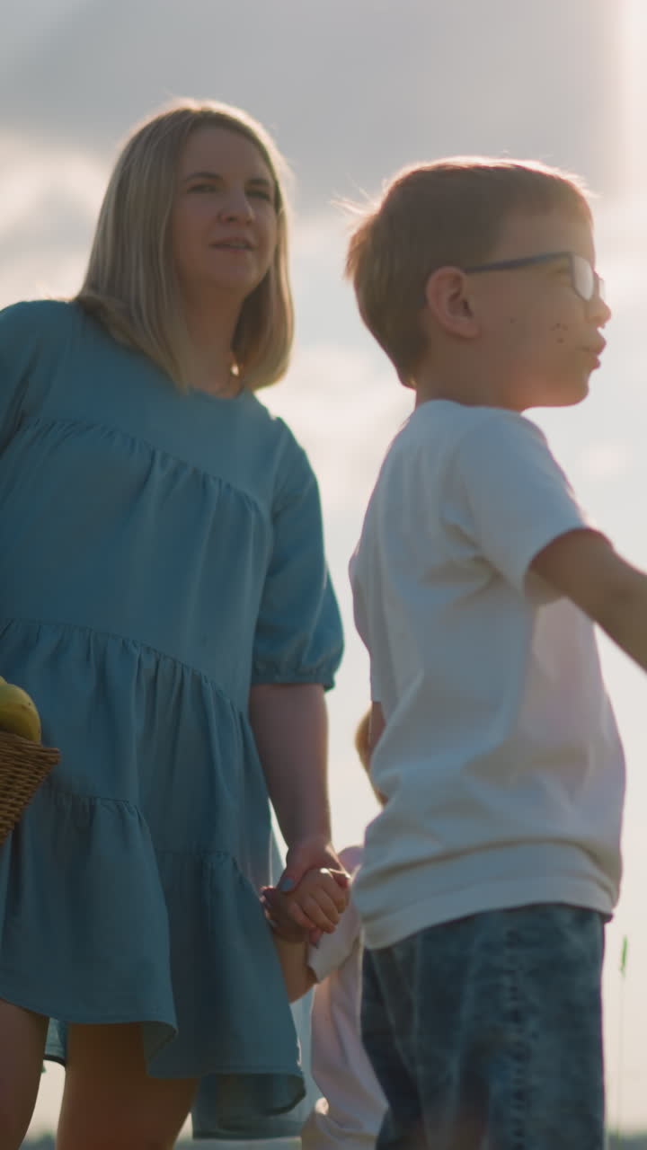 un niño con gafas y una camisa blanca señala emocionado hacia un lago mientras su madre en un vestido azul sostiene a su hermano menor, bajo un cielo parcialmente nublado, mostrando un momento de unión familiar