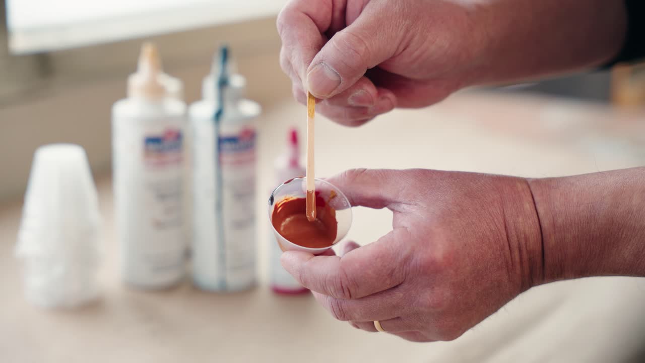 Hands display cup and stir stick of brown paint with blurred pigment bottles behind