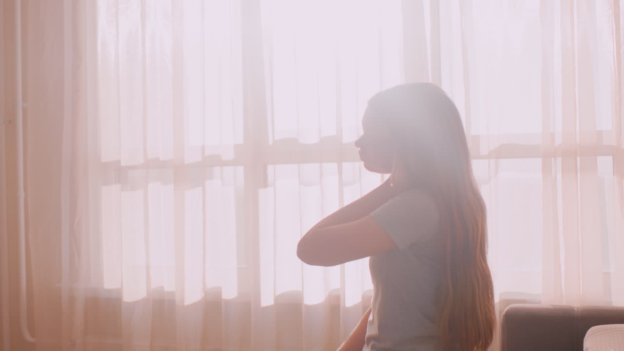Close up side view of independent girl seated near bright window gently stretching neck with hand due to tension or stiffness, expressing discomfort and care in soft sunlight through sheer curtain