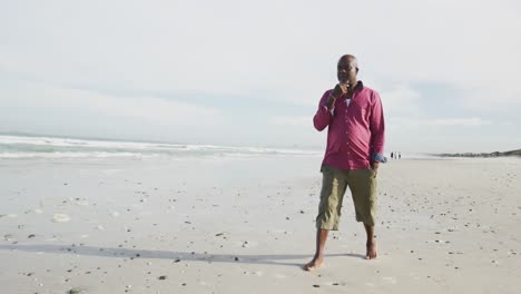 african american senior man walking on a beach rubbing his chin and looking at the sea