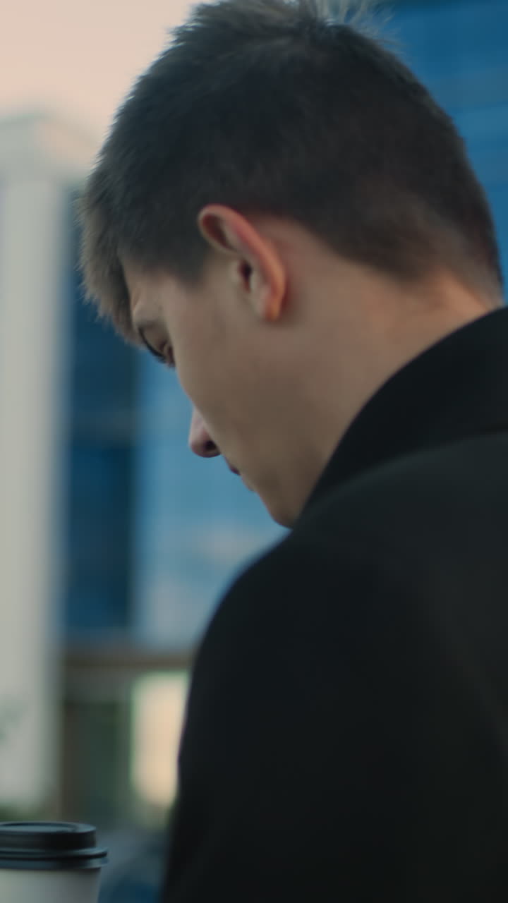 검은 코트를 입은 남자가 뒷면에 흐릿한 초록색과 현대적인 유리 건물을 특징으로하는 야외 도시 환경에서 차를 마시면서 전화에 집중하고 있습니다.