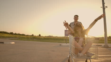 Side-view-of-a-young-man-and-woman-having-fun-outdoors-on-shopping-trolleys.-Multiethnic-young-people-racing-on-shopping-carts.-On-the-parking-zone-with-their