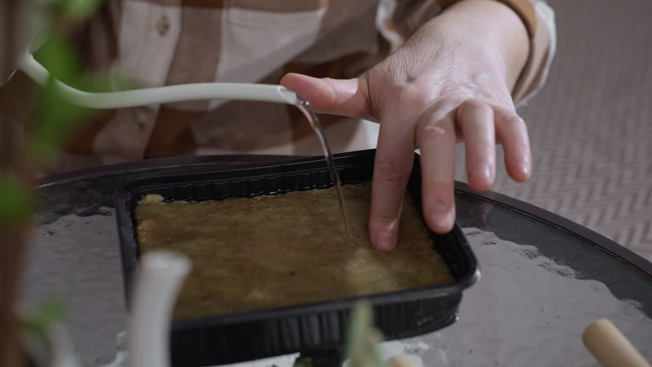 Close up of green thumbed caretaker touching surface of seed medium with fingertip while pouring water from slender spout watering can into plastic container on glass table