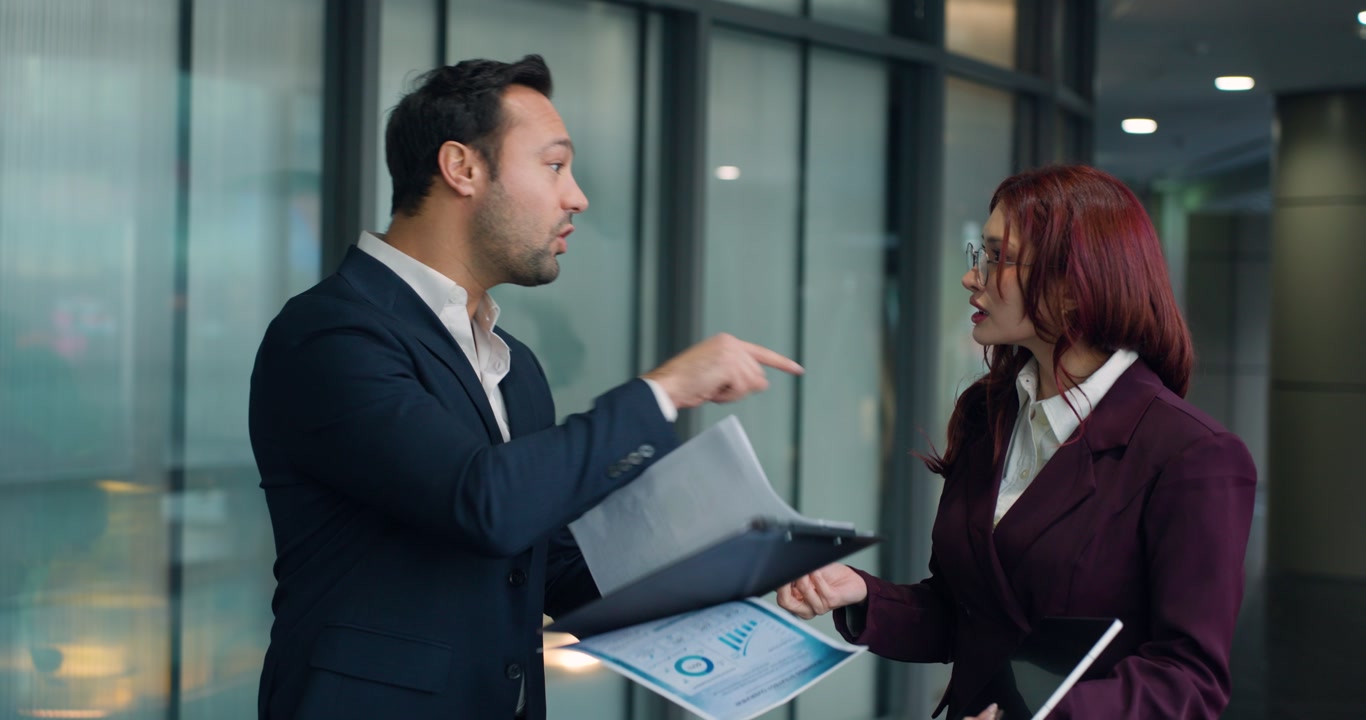 Dissatisfied businessman blames woman colleague for bad project outcomes they argue intensely reviewing documents clipboard tablet in modern corporate hallway slow motion