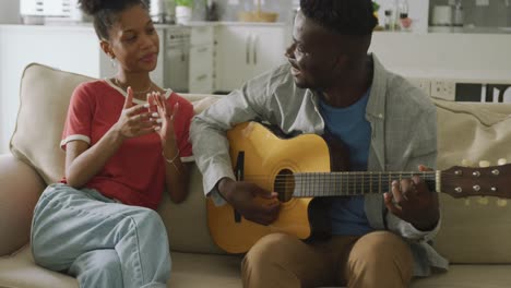 Video-of-happy-african-american-couple-sitting-on-sofa,-singing-and-playing-guitar