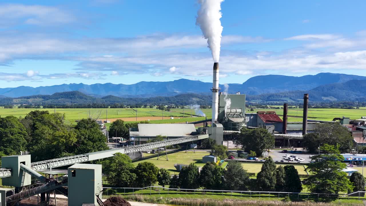 Aerial footage of a sugar mill with smoke stacks, lush greenery, and mountains under clear blue skies