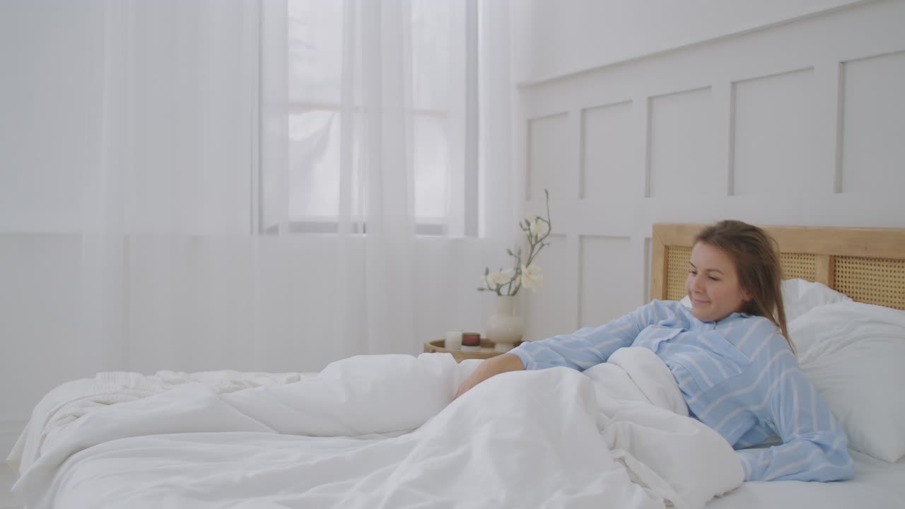 прекрасная брюнетка медленно просыпается по утрам, растягивается и встает с постели. солнце светит на нее из большого окна. счастливая молодая девушка приветствует новый день.