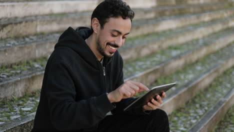 Side-view-of-excited-young-Arabic-handsome-man-with-dark-curly-hair-and-beard-in-black-hoodie-sitting-on-stairs-outside