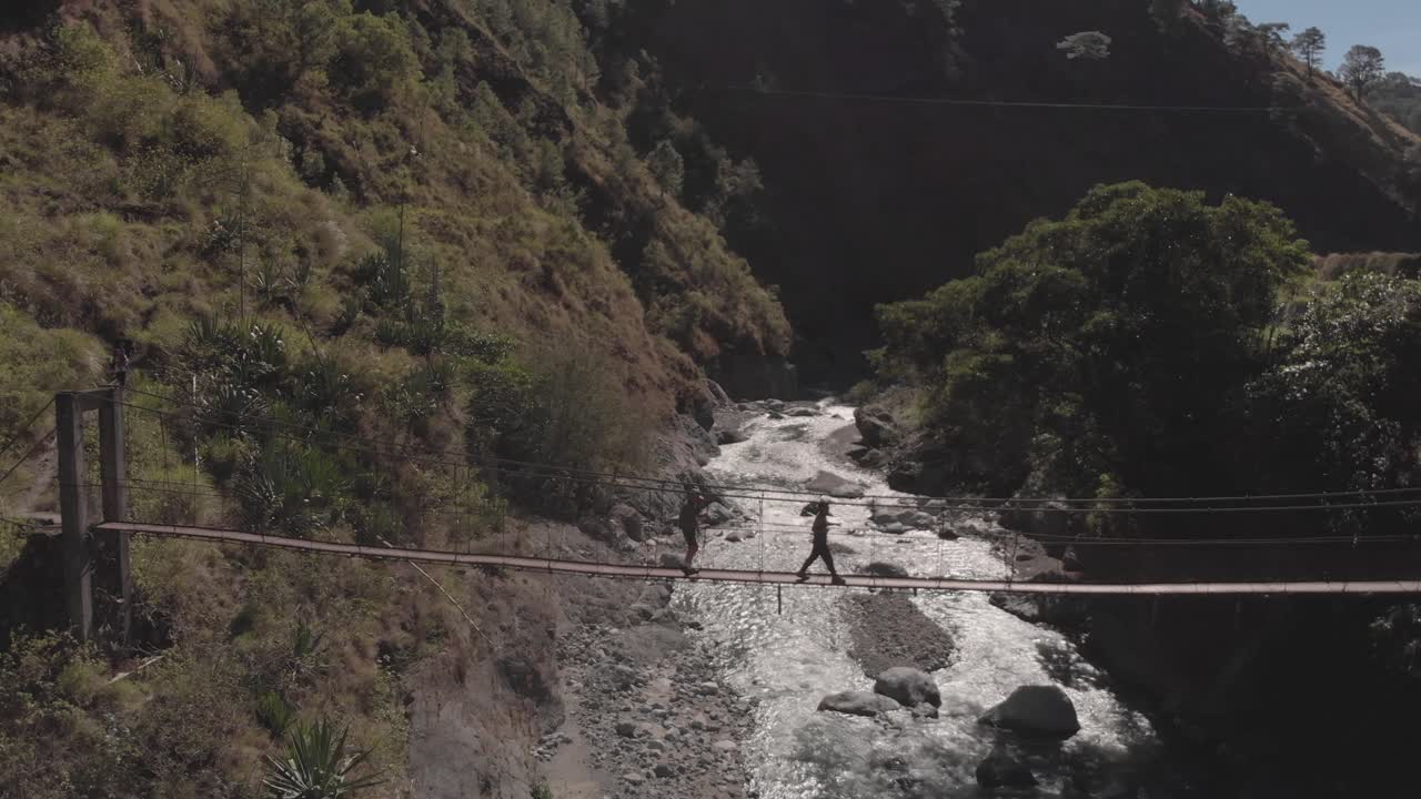 two men hiking walking across a metal wire suspension bridge spanning across a river in wild forest mountain terrain surrounded by trees green light flair reversing aerial in benguet philippines
