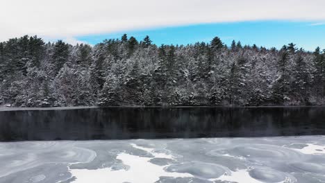 aerial push over frozen lake towards a snow covered forest shore and a strip of open water