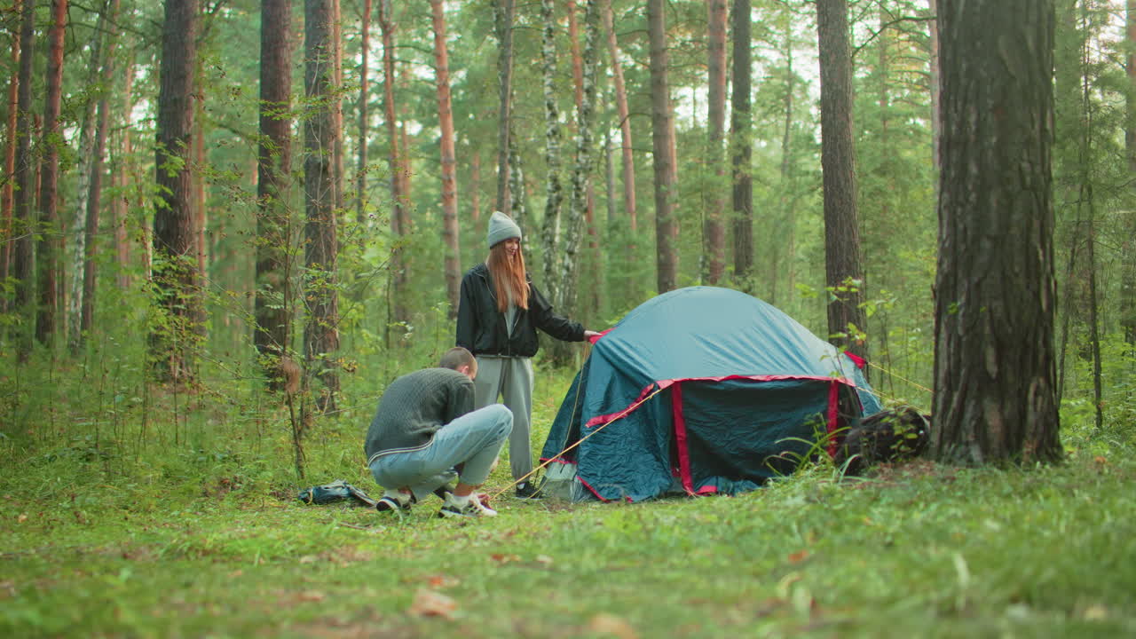 Young couple works together to secure camping tent in forest as woman holds fabric steady while man crouches down to peg yellow rope into ground surrounded by trees and morning light