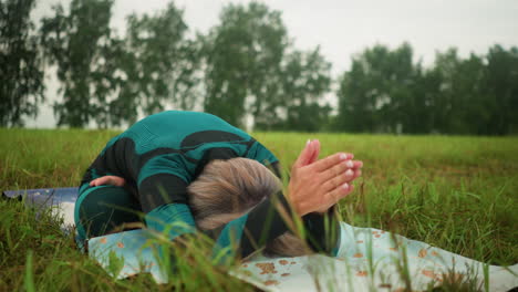 woman in green and black suit seated on yoga mat, bending forward with hands extended and head touching the mat, practicing a yoga pose outdoors in grassy field under cloudy sky