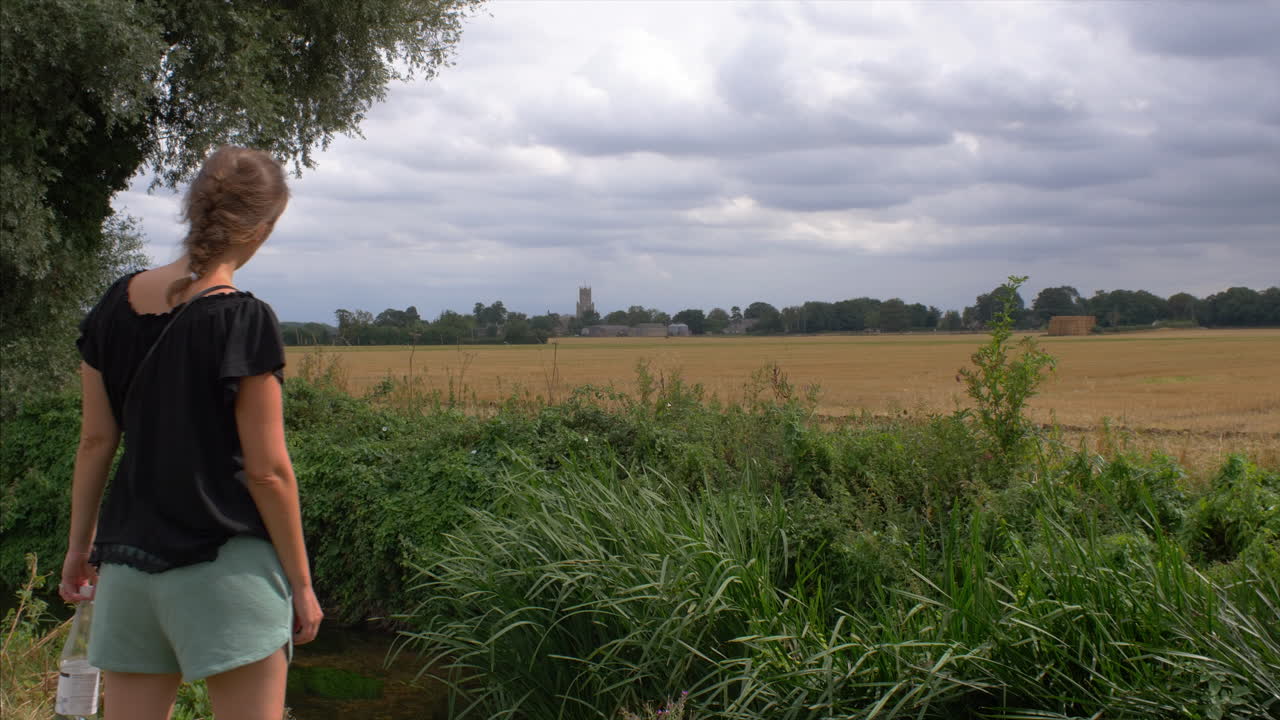 An engaged woman at a unique location in the English countryside, looking curiously over a field, with a church on the horizon and clouds in the sky.