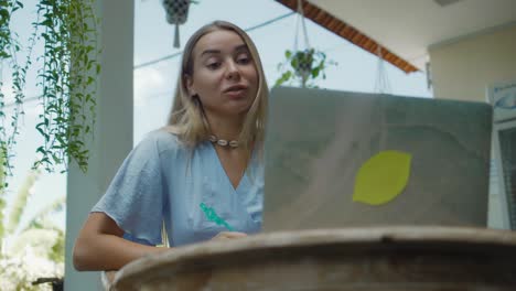 una chica en un vestido azul remotamente en línea trabajando detrás de una computadora portátil y mirando a la pantalla el patio trasero con plantas verdes