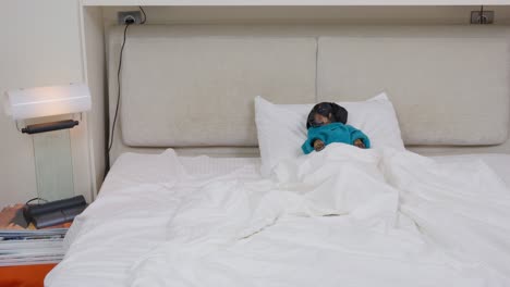 un cachorro de perrito negro vestido con una túnica de terry azul se acuesta cómodamente en la almohada de la cama bajo una manta blanca y cálida y huye