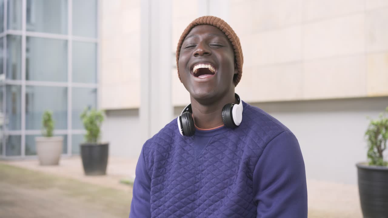 Afroamerikansk man med mössa och hörlurar reagerar på ett skämt utanför bilden och skrattar på en innergård.