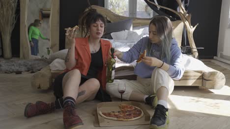 two happy girls eating pizza on the floor while their two friends sitting on the bed and one of them recording with video camera