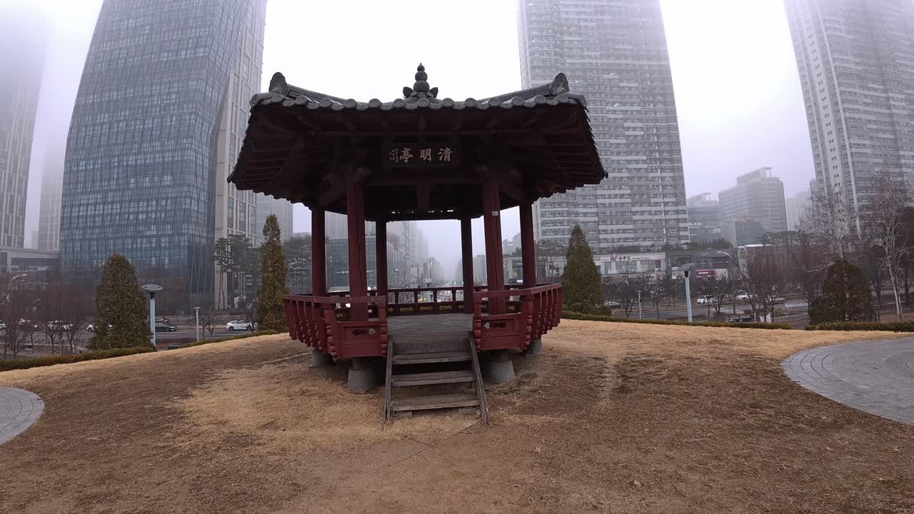 Songdo Central Park - Traditional Gazebo Backdropped By Modern High-rise Buildings In Fog - Incheon, South Korea. pullback shot