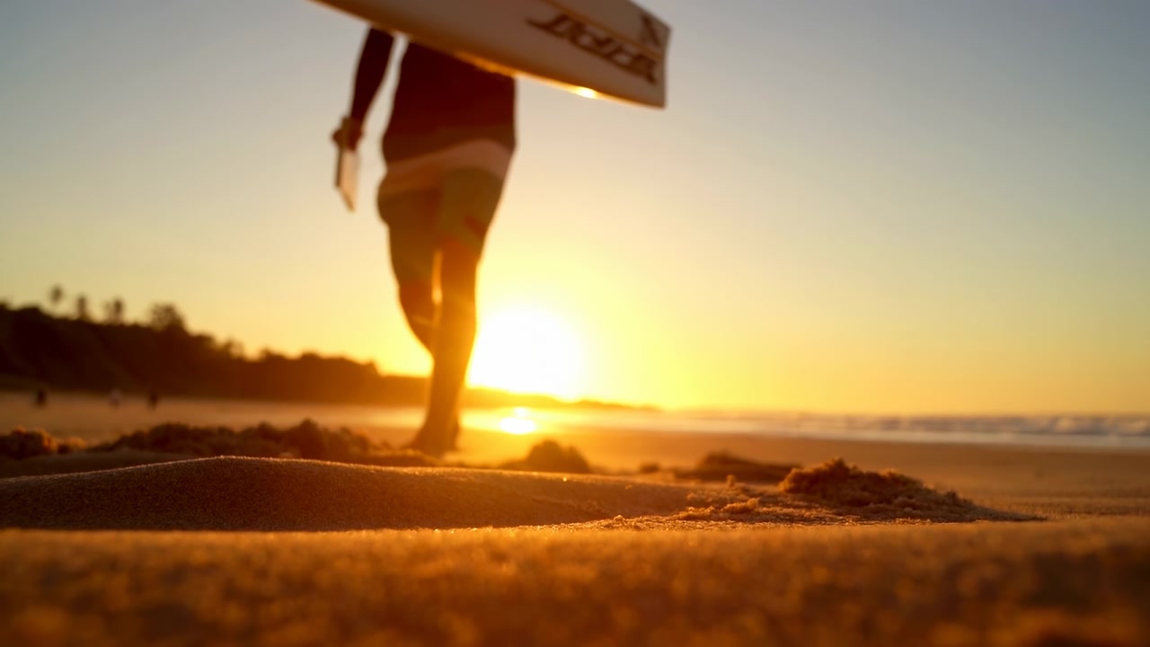 Low-angle view follows bare feet crossing warm textured sand while surfboard passes overhead, with dangling leash visible against bright sunset, calm sea horizon, and golden coastal light.