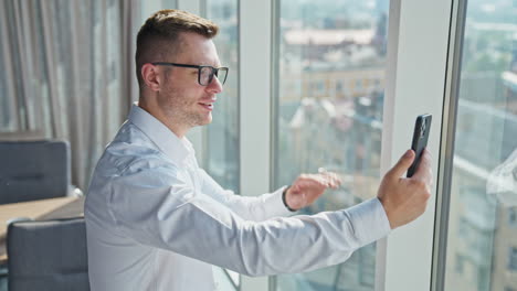 mid-aged handsome man in white shirt and glasses in a sunny office. businessman having video chat on his smartphone. side view.