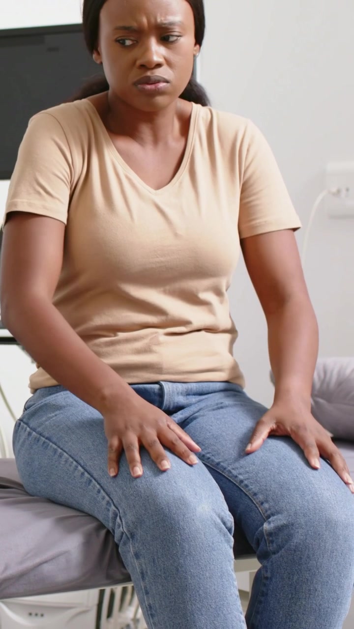 Vertical video: Feeling pain woman rubbing abdomen pressing thighs bracing on exam table in tan tee