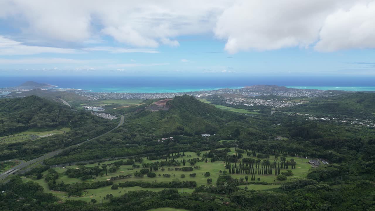 Wide static drone shot of Big Island's coastline, city, forest, and mountains under a partly cloudy sky.
