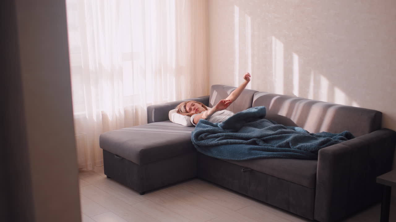 House resident stretches on couch under blanket, slightly pushing it down while moving leg as morning sunlight streams through curtains, cozy domestic atmosphere with warmth
