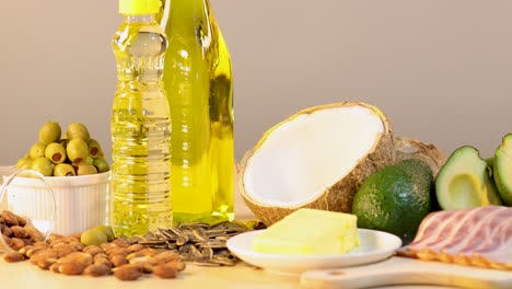 a variety of high-fat foods including olive oil, coconut, avocado, butter, bacon, almonds, and olives arranged on a wooden surface with soft studio lighting and smooth camera pan