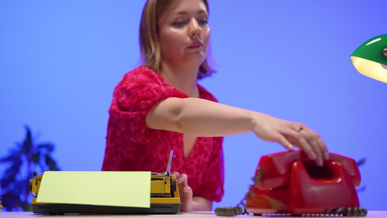 A woman in a red floral dress reaches toward a red rotary phone on her retro desk, which also features a yellow typewriter and soft studio lighting with a blue backdrop and green banker’s lamp