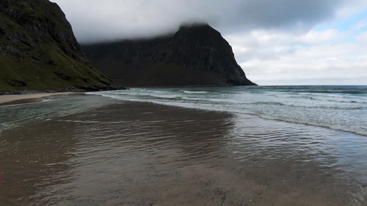Flyver over havet og bølgerne, der brækker ved en strand med udsigt til skyindhyllede bjerge og stranden ved foden af bjerget i slowmotion