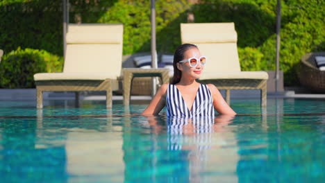 while standing in waist-high pool water an attractive young woman adjusts her sunglasses