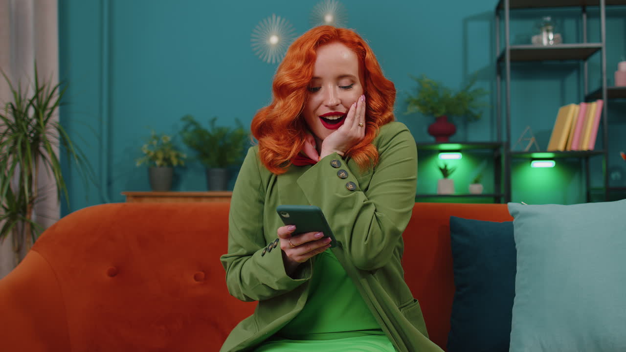 Giovane ragazza dai capelli rossi a casa sul divano usa lo smartphone scrive la navigazione dice wow sì ha scoperto grande grande vittoria