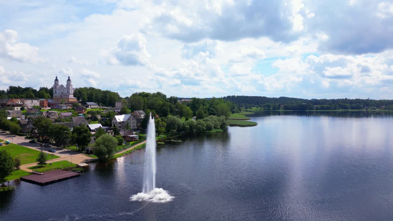 Wide aerial view of large fountain spraying over lake near historic church and village edge. Shot at Zarasai, Lithuania