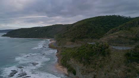 Reveal-shot-of-Australia's-Great-Ocean-Road-coastal-highway-on-cloudy-overcast-day-with-crashing-surf-waves-in-Victoria