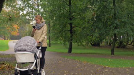 Frau-Benutzt-Handy-Beim-Spazierengehen-Mit-Baby-Im-Herbstpark